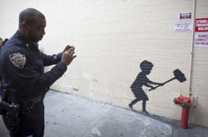 New York City cop taking a picture with his phone of Banksy graffiti art.